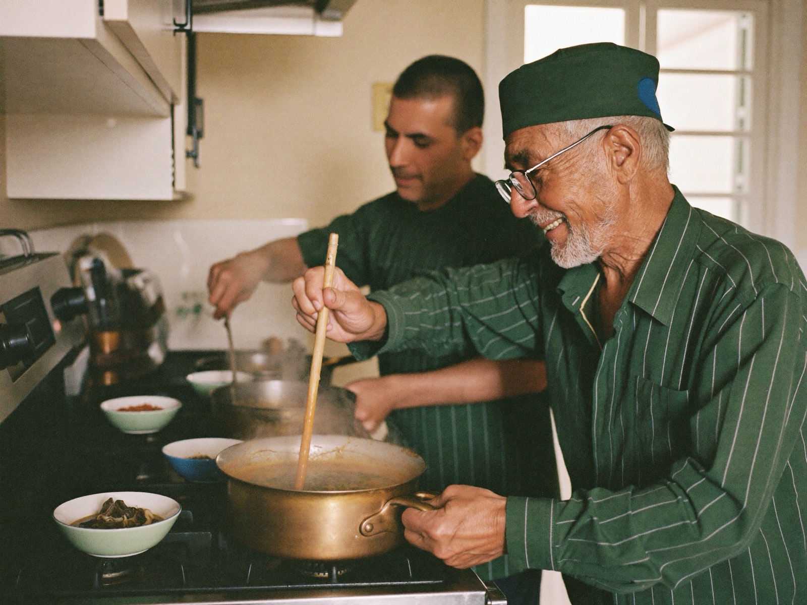An older man and his regular carer cooking together at the stove in a warm family kitchen.