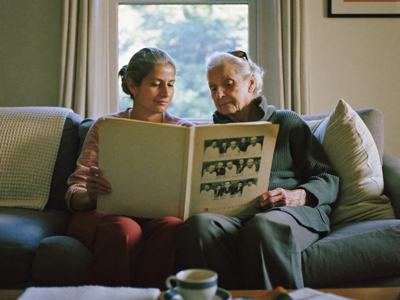 A daughter sitting close beside her elderly mother on a sofa, looking through a family photo album.