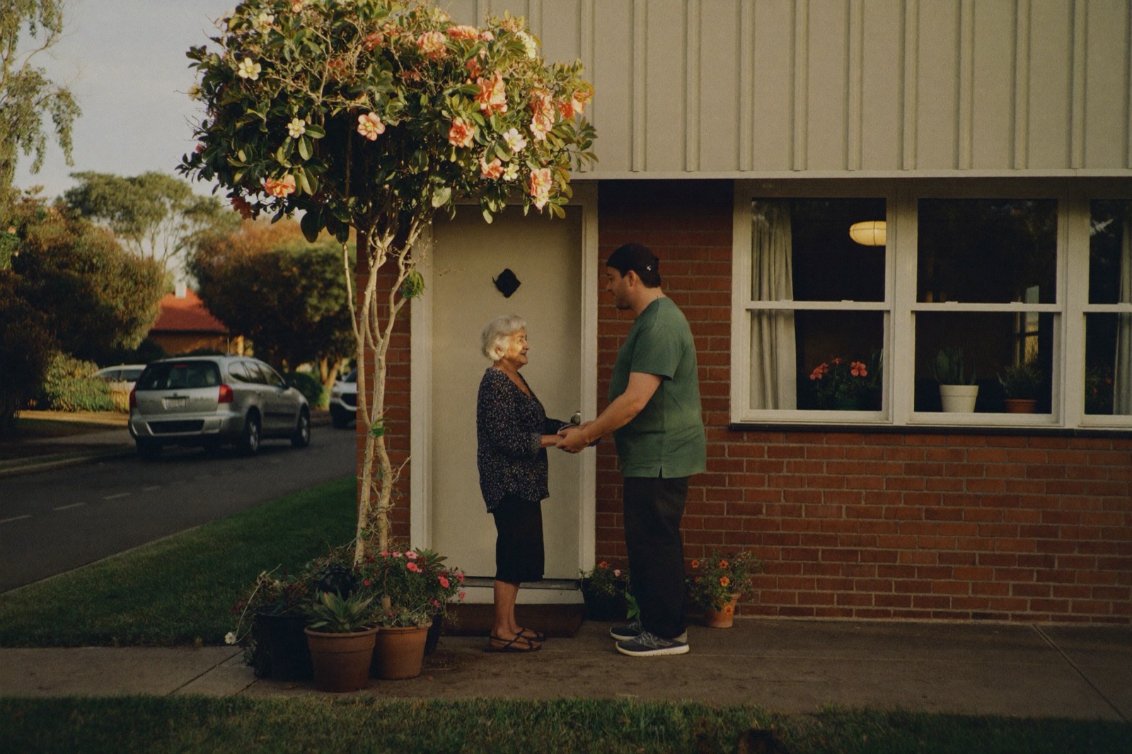 An older woman warmly greeting her regular carer at the front door of her red-brick home.