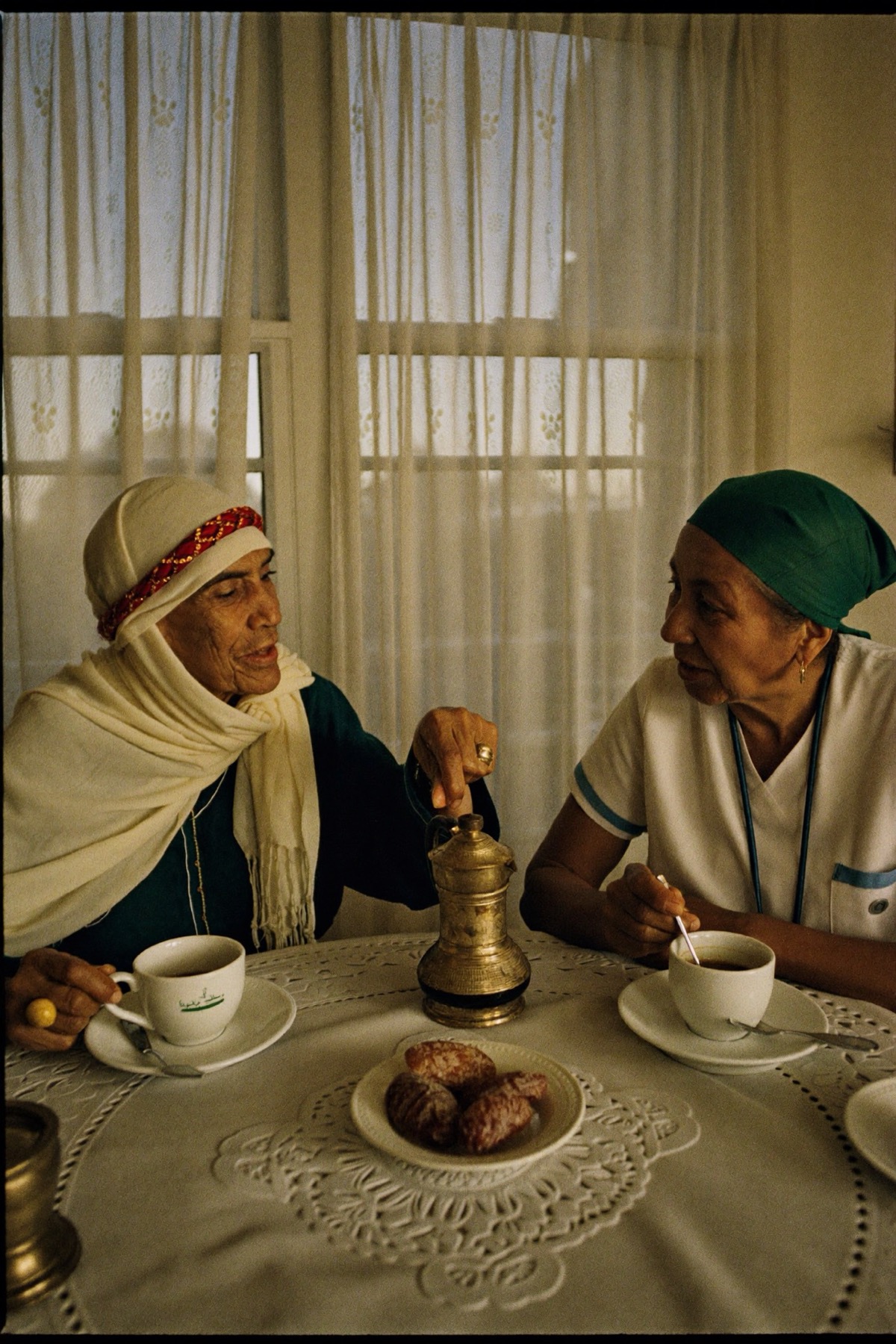 An older woman sharing Arabic coffee with her bilingual carer at a lace-covered table.