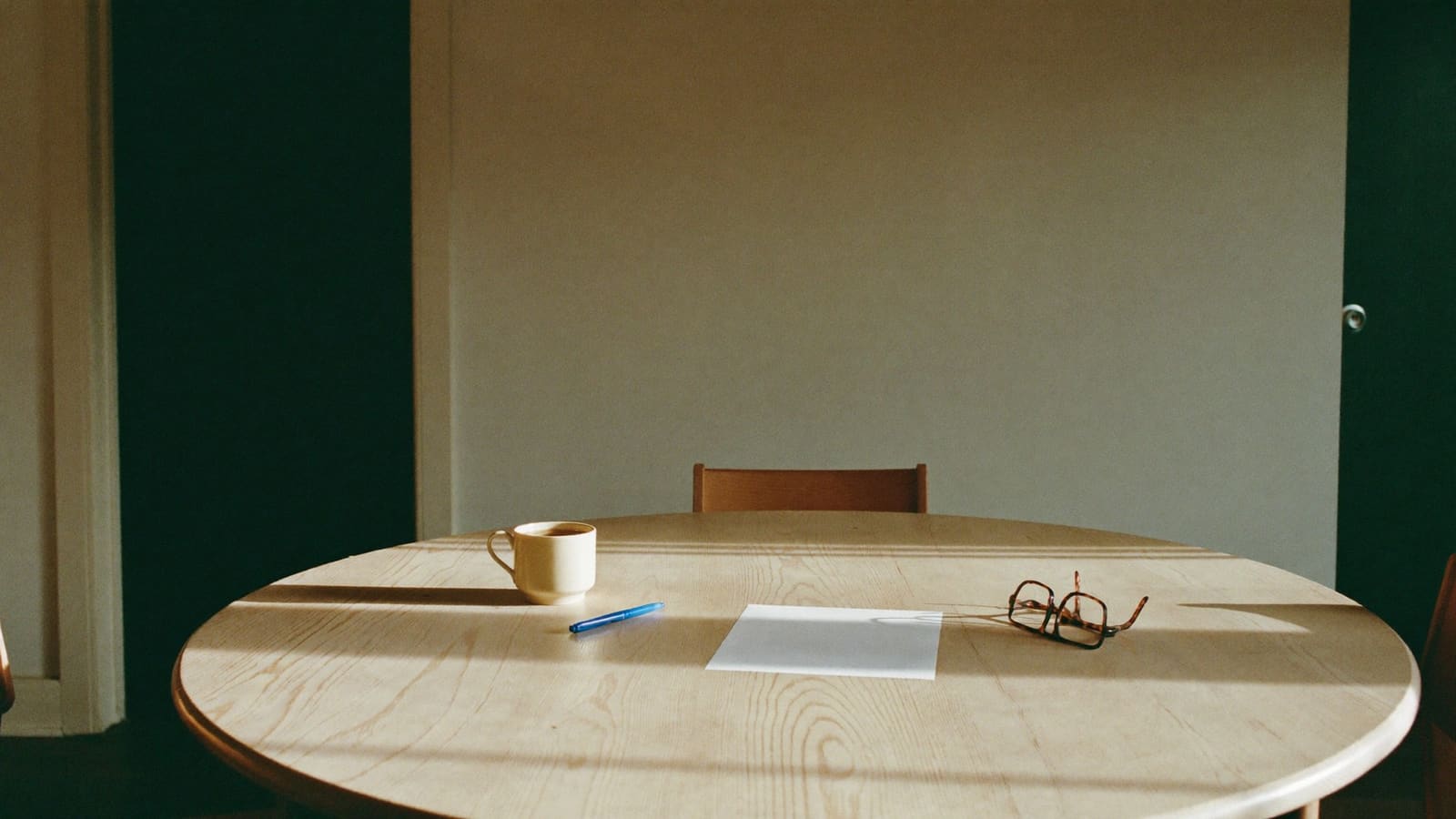 A cup of tea, folded reading glasses and a pen on a timber dining table, with a single blank page face down beside them.