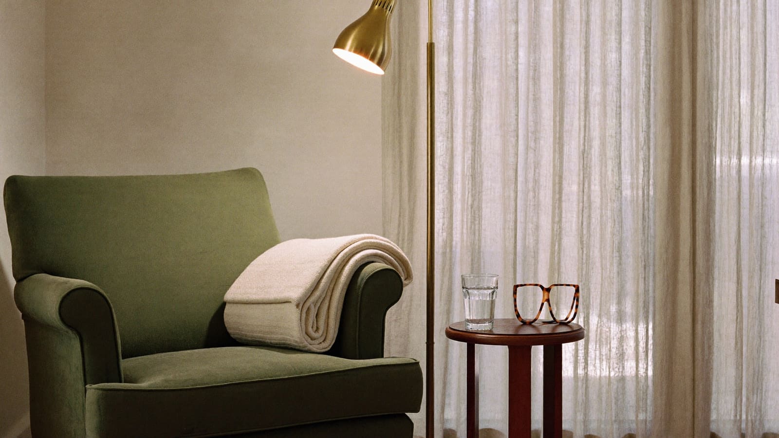 A sage green armchair with a folded cream rug over one arm, a brass floor lamp switched on, and a small timber side table holding a glass of water and a pair of tortoiseshell reading glasses.