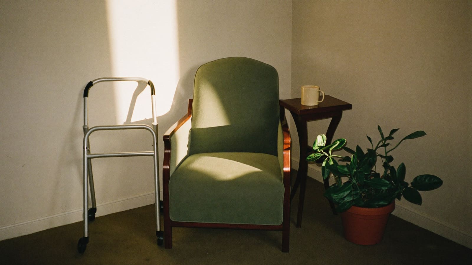 A lightweight walking frame standing beside an olive green armchair in a sunlit living room.