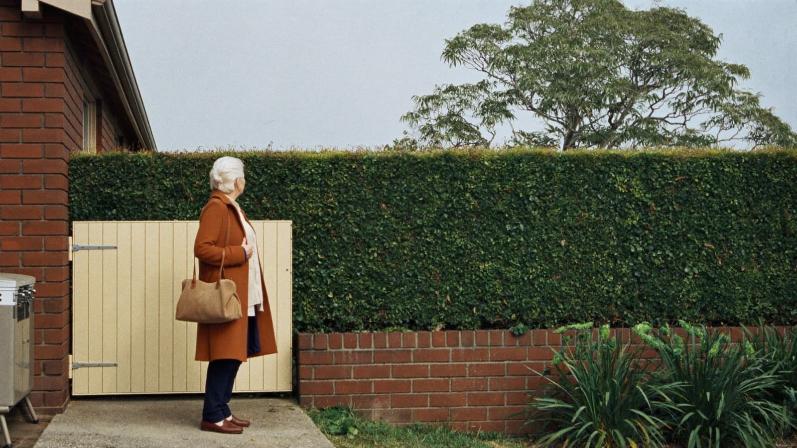 An older woman in a coat with a handbag waiting at the front gate of her brick house.