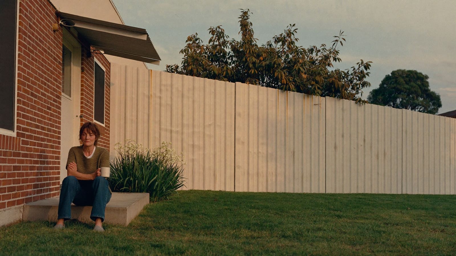 A woman in her fifties sitting alone on the back step of a suburban house with a mug of coffee, shoulders loose.