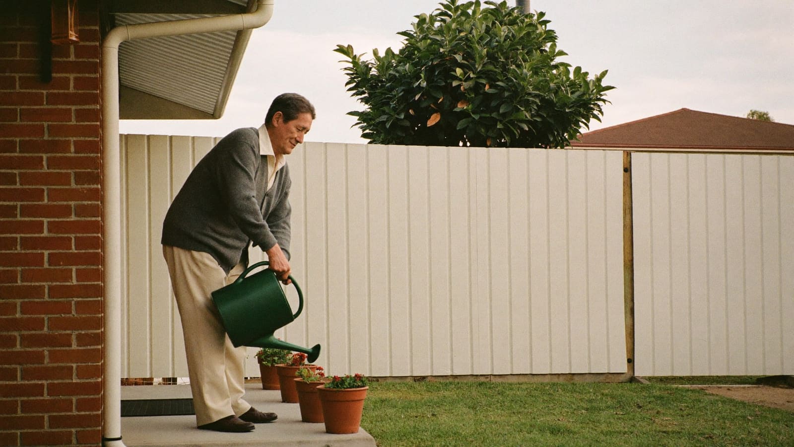 An older man watering terracotta pots on the concrete back step of his home.