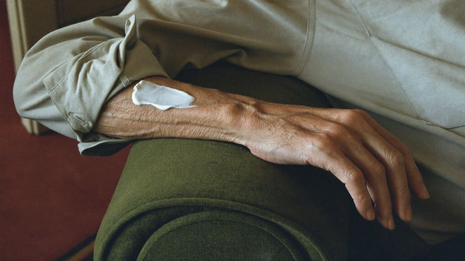 The bare forearm and hand of an older man resting along the padded arm of a worn olive green armchair, with a small amount of white moisturiser freshly applied to the thin, softly lined skin below the rolled shirt cuff.