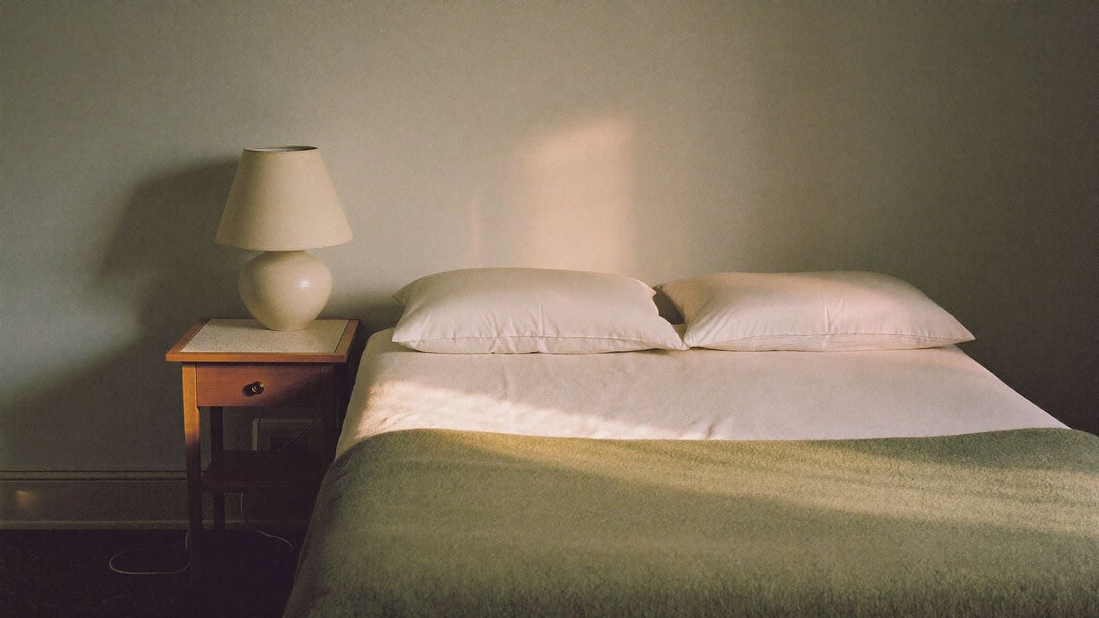 A neatly made bed with cream bedding and a folded sage green blanket in a quiet sunlit bedroom.