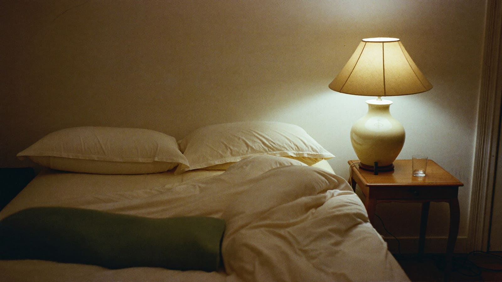 A bedside lamp lit in a dark bedroom with the covers turned back on one side of the bed.
