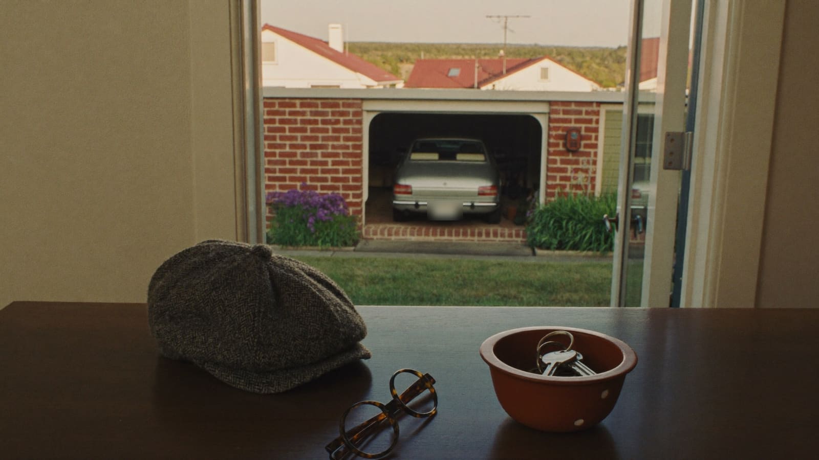 Car keys in a small terracotta bowl on a hallway table beside a tweed cap and glasses, with an older sedan parked in a brick carport outside.
