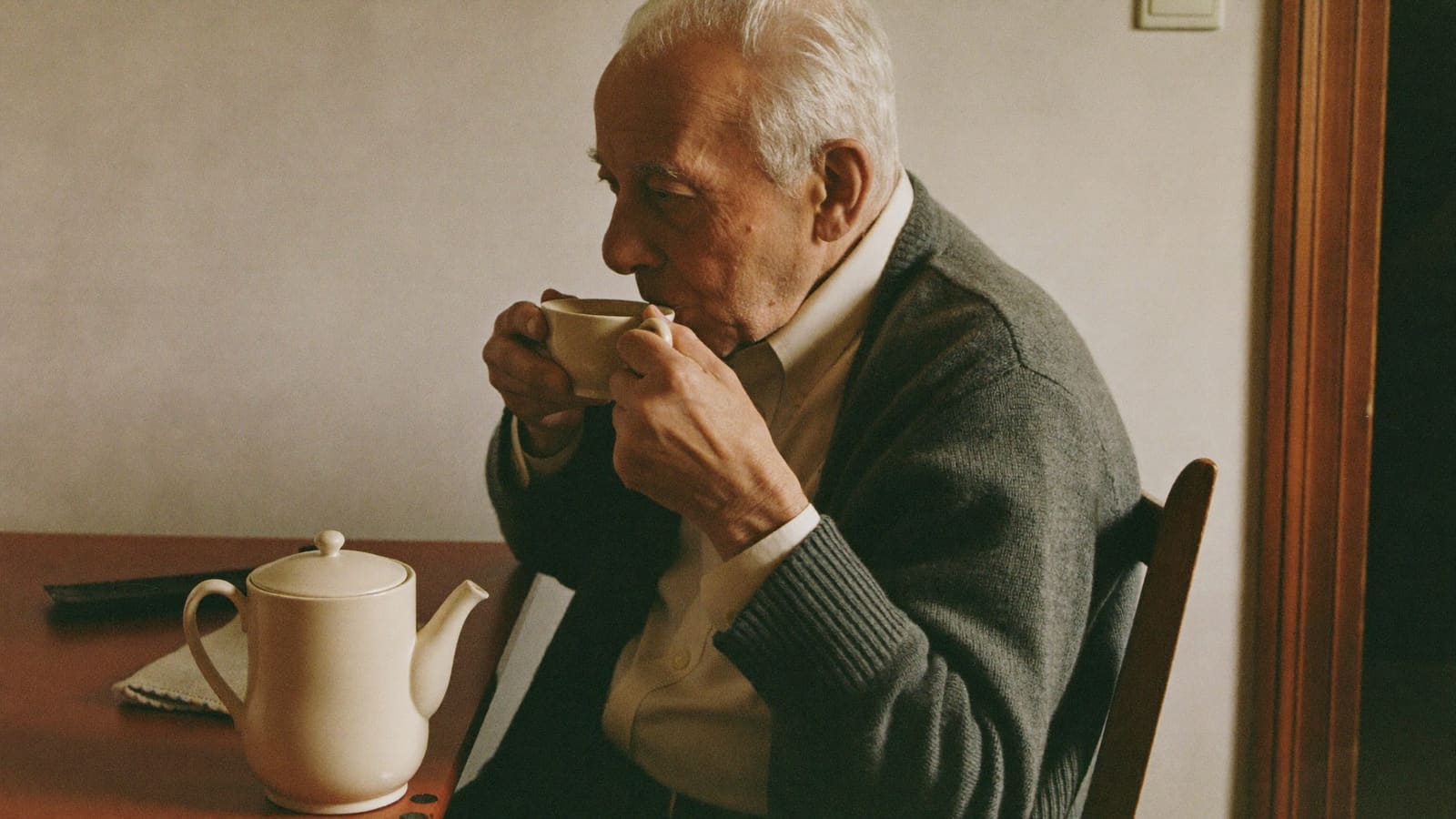 An older man taking a careful sip of tea from a cup held in both hands at his kitchen table.