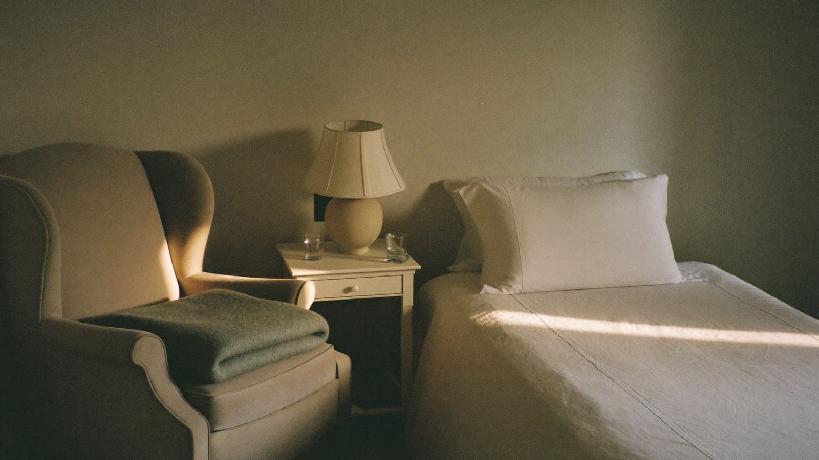 An armchair pulled up close alongside a neatly made bed in a quiet bedroom.