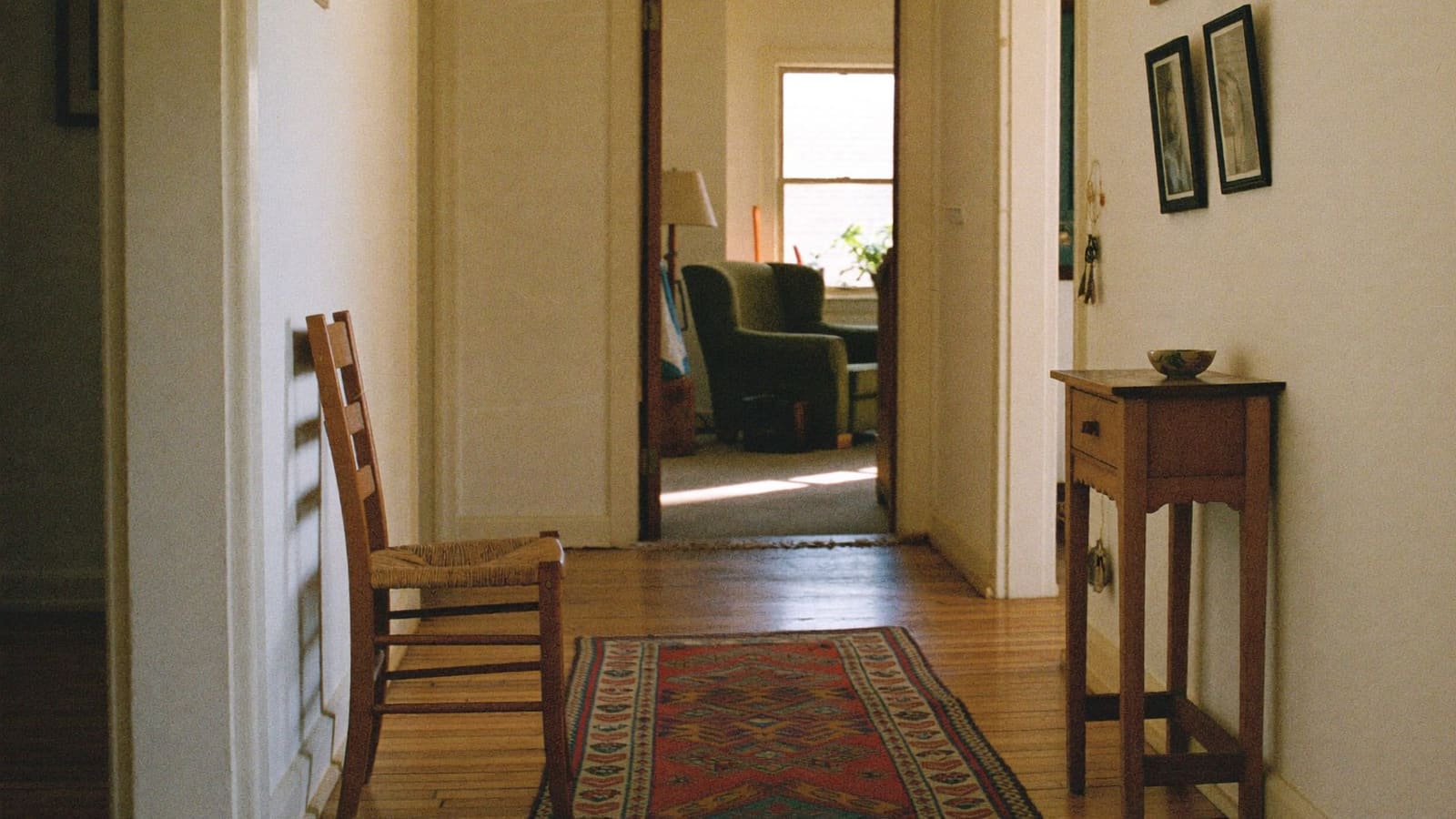 A single plain timber chair with a woven seat placed against the wall halfway along the hallway of an ordinary brick home, with framed family photographs opposite and an open doorway to a sunlit living room at the far end.