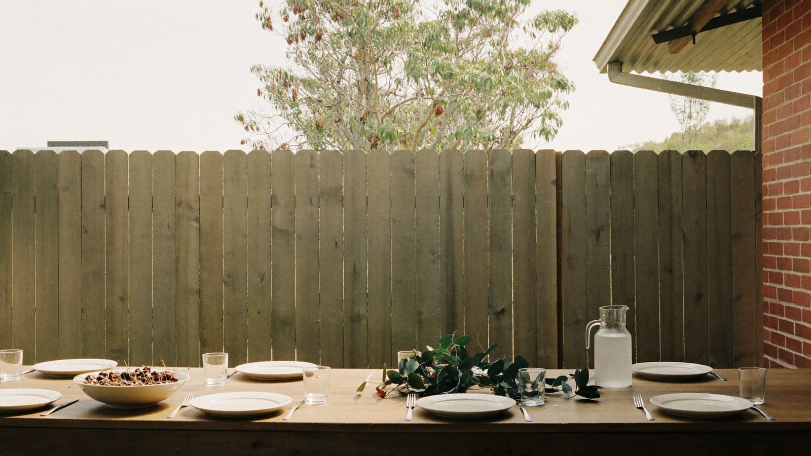 A long timber table on a shaded back veranda set with white plates, silver cutlery, a bowl of cherries, a jug of iced water and loose sprigs of eucalyptus, with a tall paling fence and a gum tree behind it.