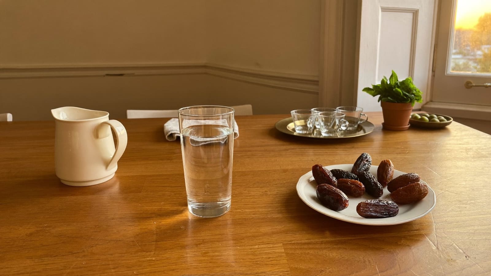 A kitchen table laid at sunset with a tall glass of water, a saucer of dates, a brass tray of small tea glasses and a pot of mint.