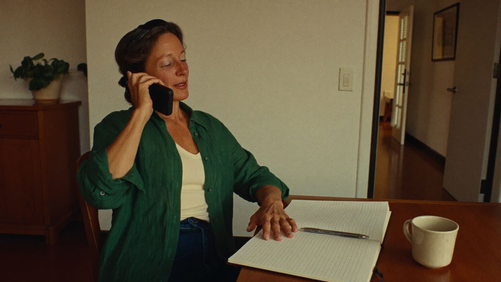 A woman in her fifties sitting alone at her dining table with a notebook and a mug, thinking.