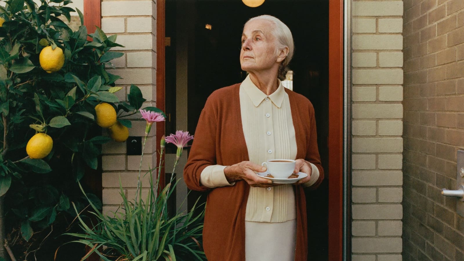 An older woman standing in her own front doorway with a cup of tea, looking out over her garden.