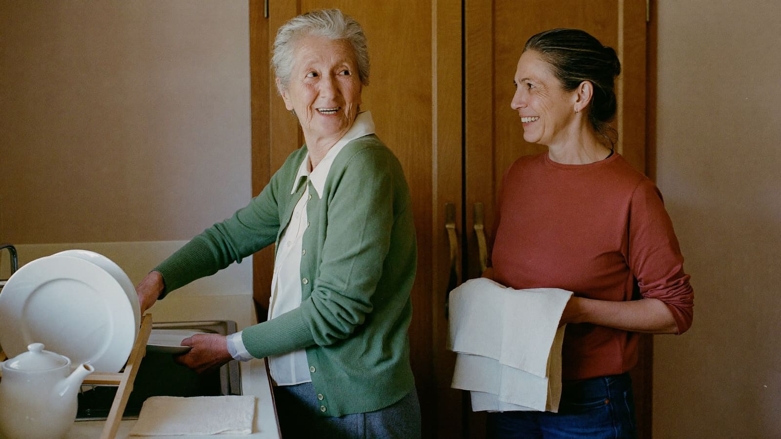 An older woman and her adult daughter drying up together at a kitchen bench, mid-conversation.