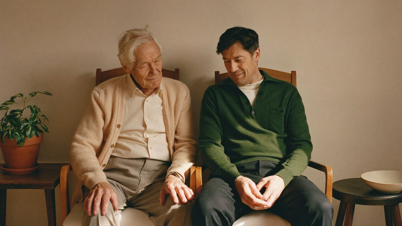 An older man and his adult son sitting close together on adjacent chairs in a sunlit living room.