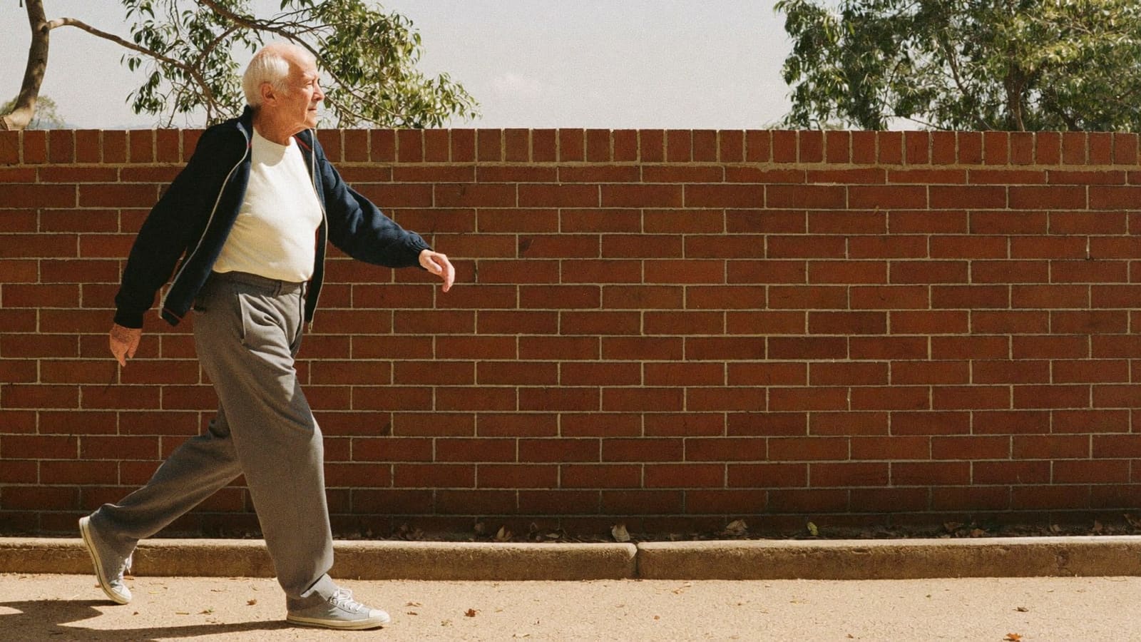 An older man walking alone at a good steady pace along a suburban footpath on a bright morning.