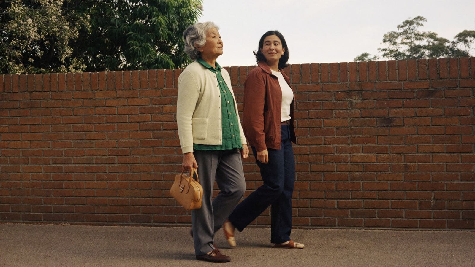 An older woman and a support worker walking side by side along a suburban footpath on a weekday morning.