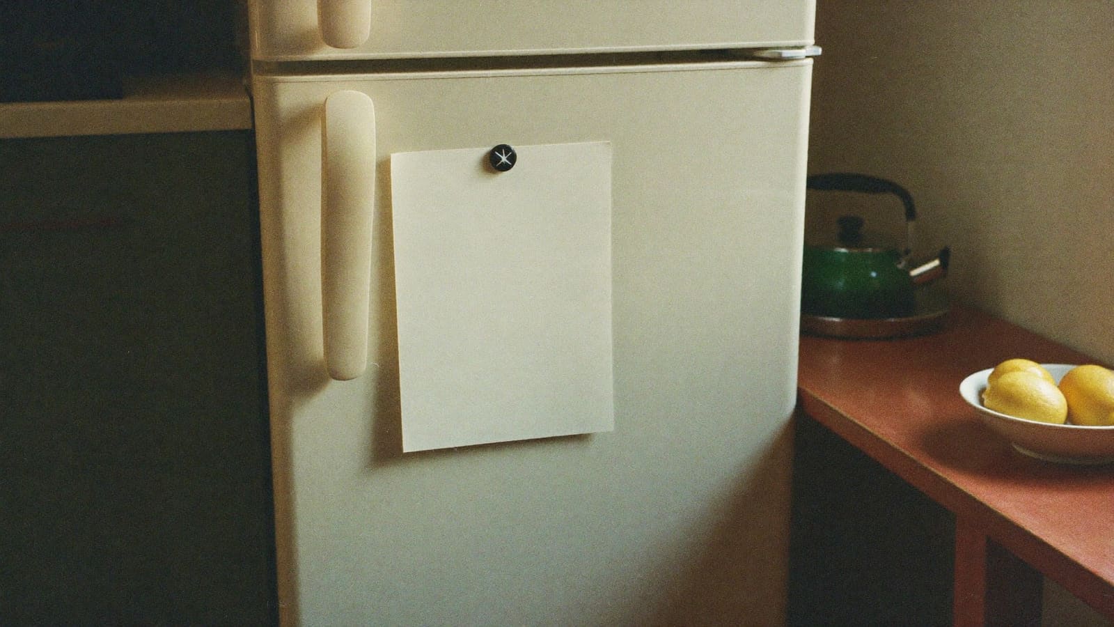 A single sheet of paper held by a plain magnet on a cream fridge door in a warm home kitchen, a green kettle on the bench beside it.