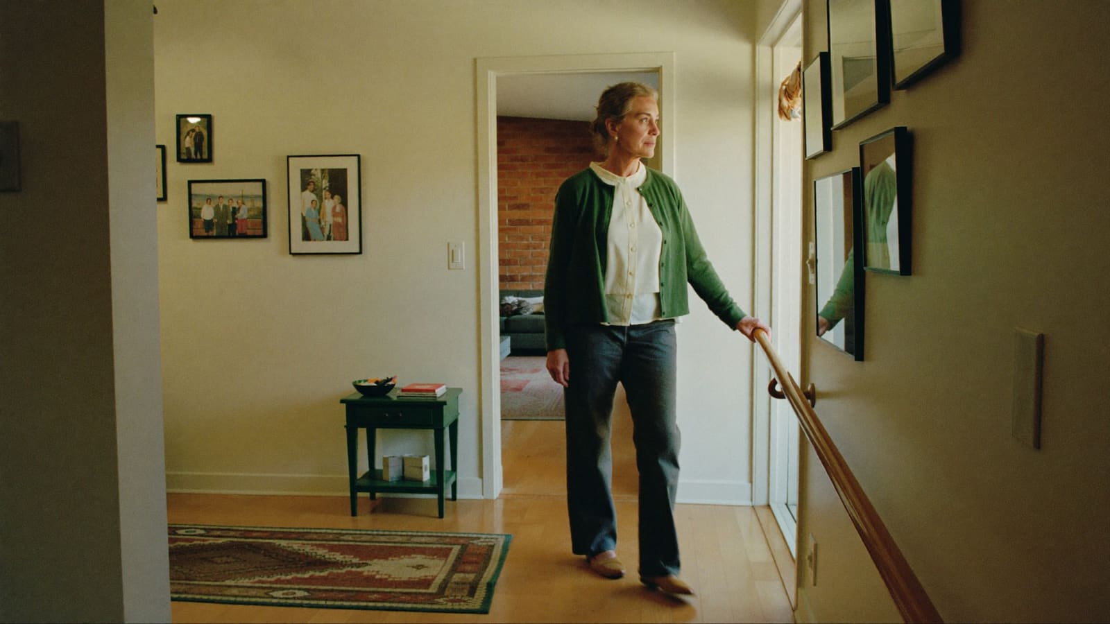 An older woman walking through a bright hallway at home, one hand resting on a timber rail.
