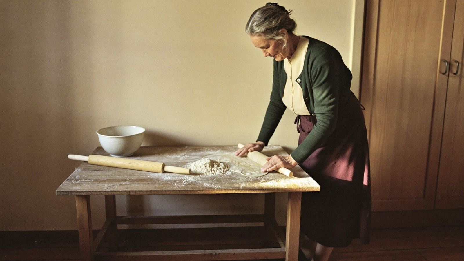 An older woman in a deep green cardigan and apron rolling out dough at her kitchen bench, absorbed in the work.