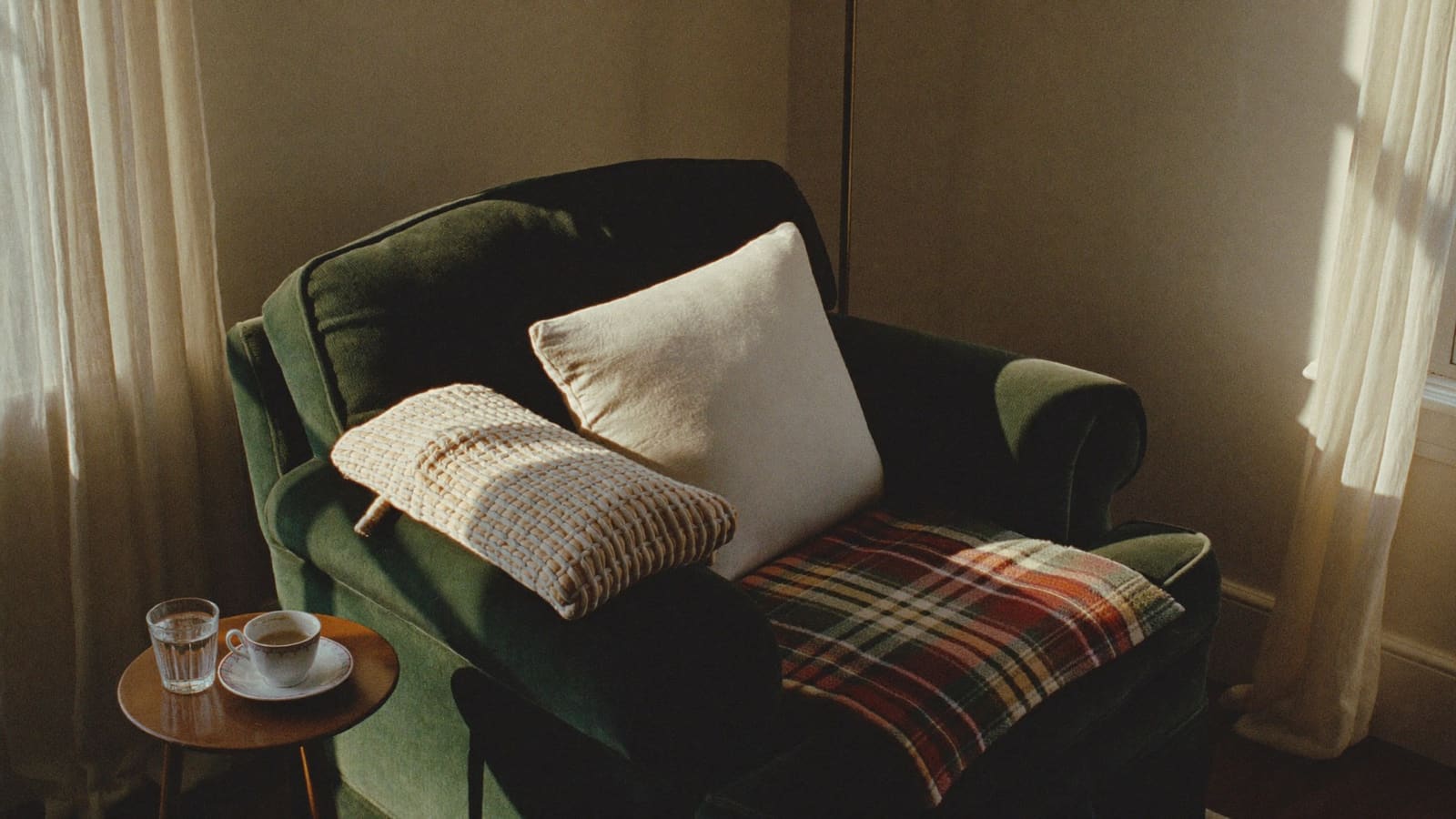 A deep green armchair in late-afternoon light with a woven wheat-bag heat pack, a cream cushion and a tartan rug, beside a small timber table with a cup of tea and a glass of water.