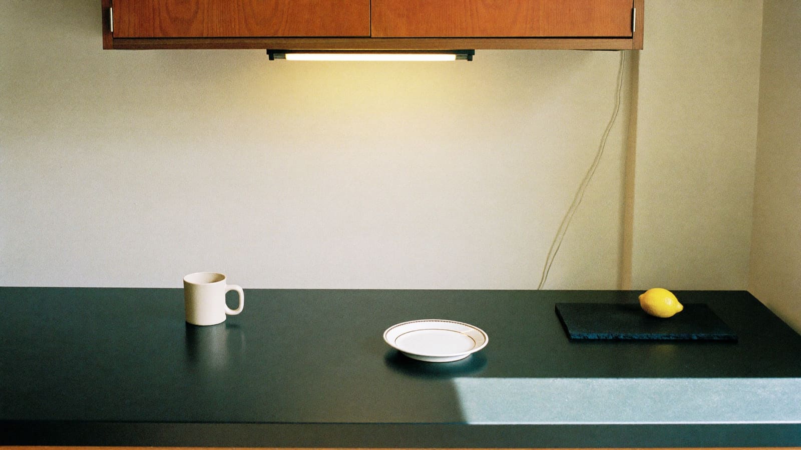 A brightly lit kitchen corner where a white mug, a white plate and a yellow lemon on a dark slate board each stand out sharply against a deep charcoal benchtop, with a strip light switched on under a timber cupboard.