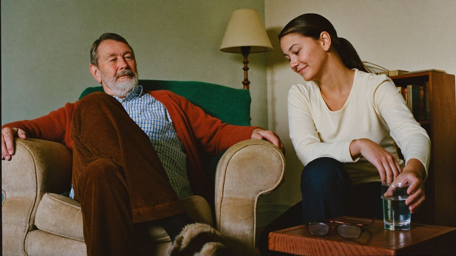 An older man settling back into his own armchair while his adult daughter sets down a glass of water.