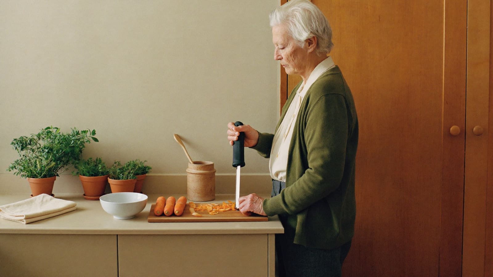 An older woman in an olive cardigan preparing carrots at her kitchen bench with a thick-handled tool held in her whole hand, her eyes down on her work.
