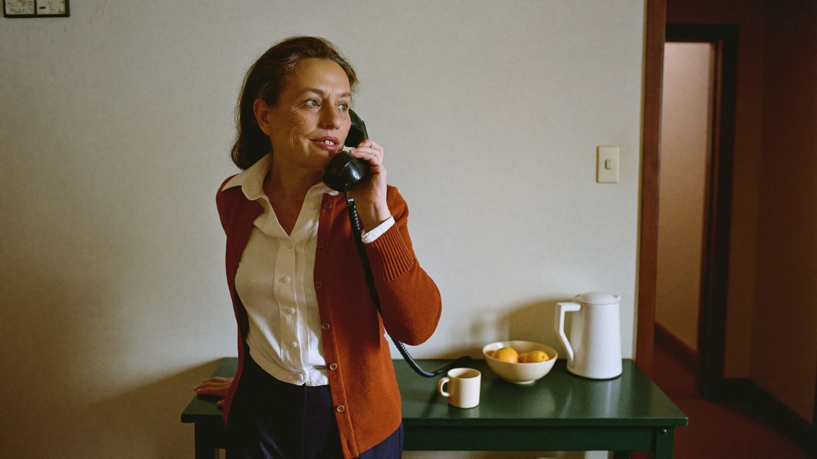 A woman in her fifties standing at a kitchen bench with a telephone handset to her ear, part way through a patient conversation.