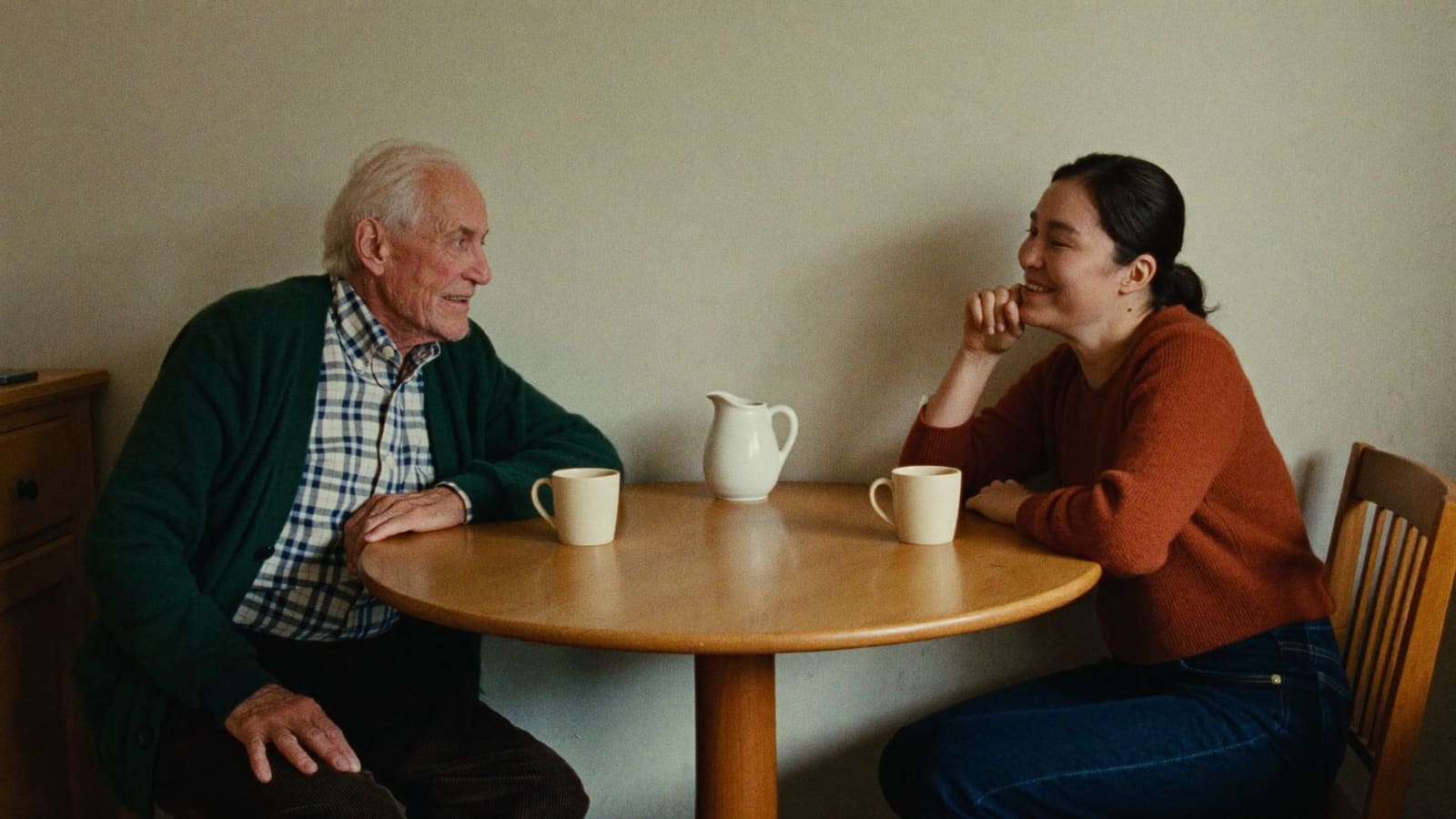 An older man and his adult daughter sitting close at the corner of a kitchen table, talking over two mugs of tea.
