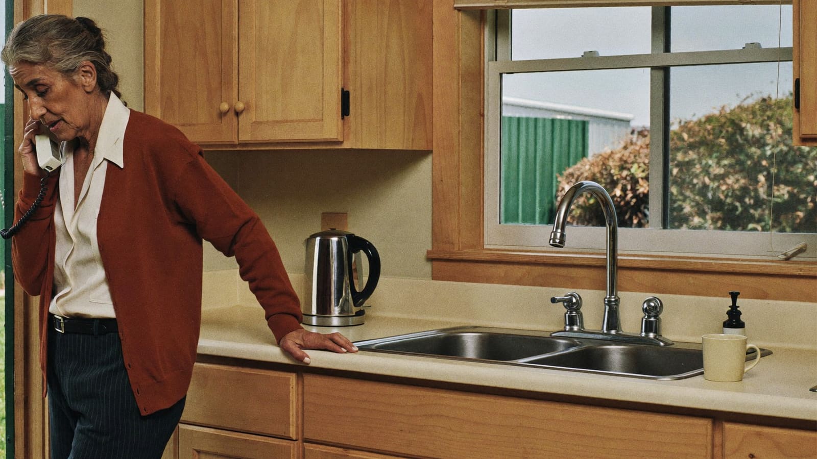 An older woman standing at her kitchen bench on the phone, one hand resting on the benchtop.