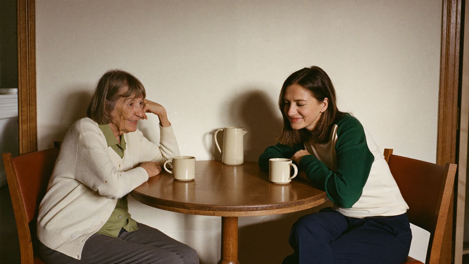 An older woman and a younger woman sitting at a round kitchen table with two mugs, talking easily.