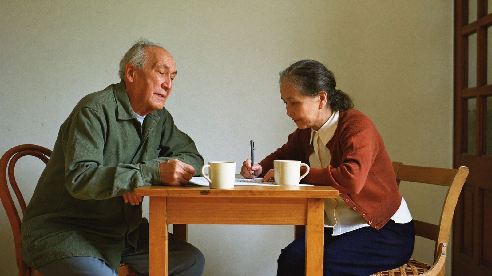 An older man and an older woman sitting close at a small kitchen table, working something out together.