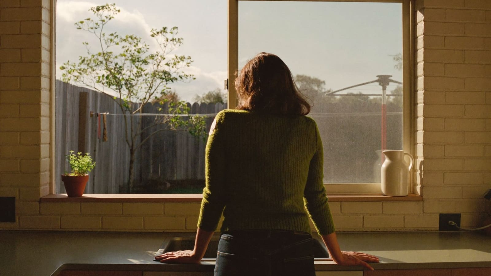 A woman in her fifties in a moss-green jumper standing at a kitchen sink, hands on the bench, looking out at the back garden.