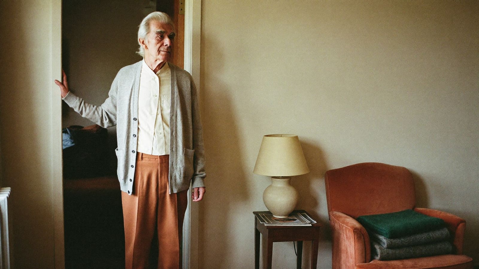 An older man standing in his living room doorway with a hand on the frame, pausing before he goes in.