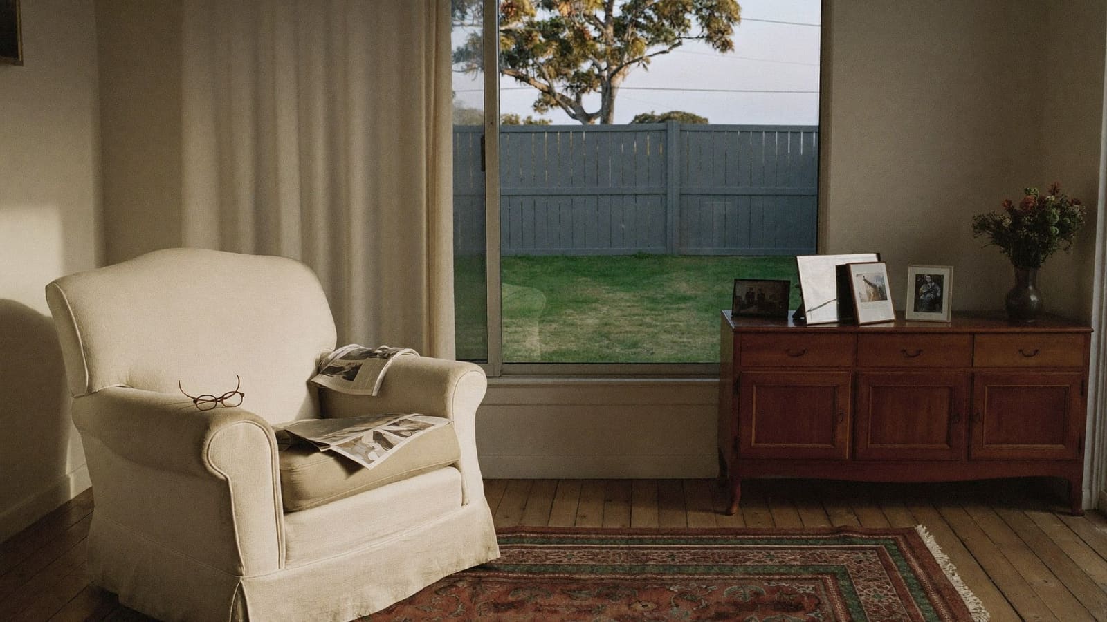The lounge room of a long-lived-in Australian home, a worn armchair by the window and a sideboard of framed family photographs.