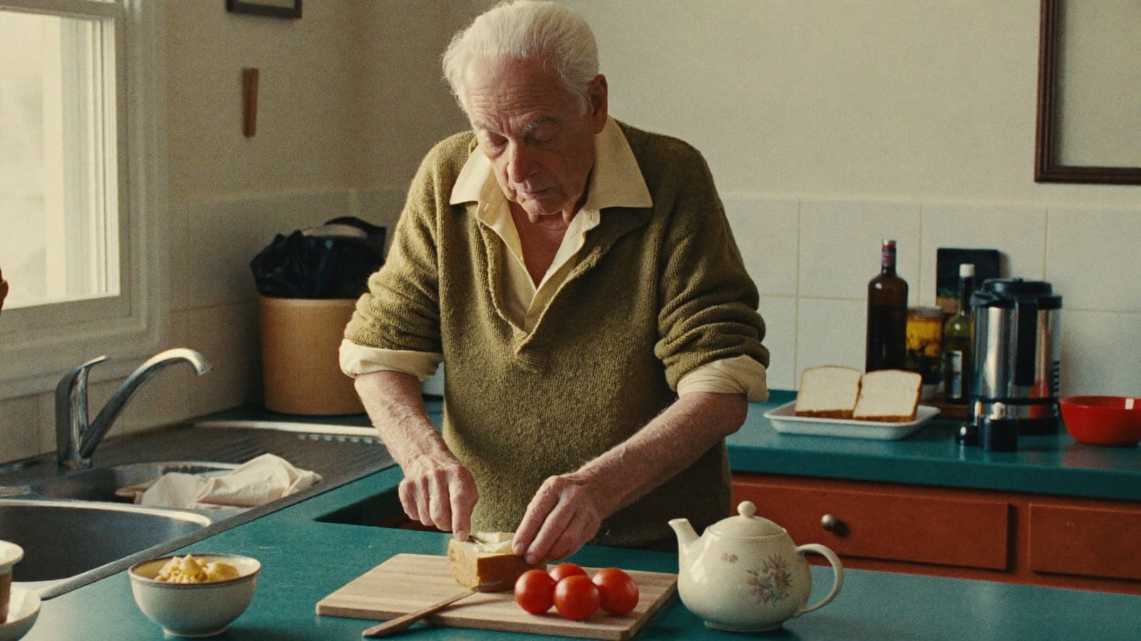 An older man making himself lunch at his own kitchen bench, sleeves pushed up, on an unhurried morning.