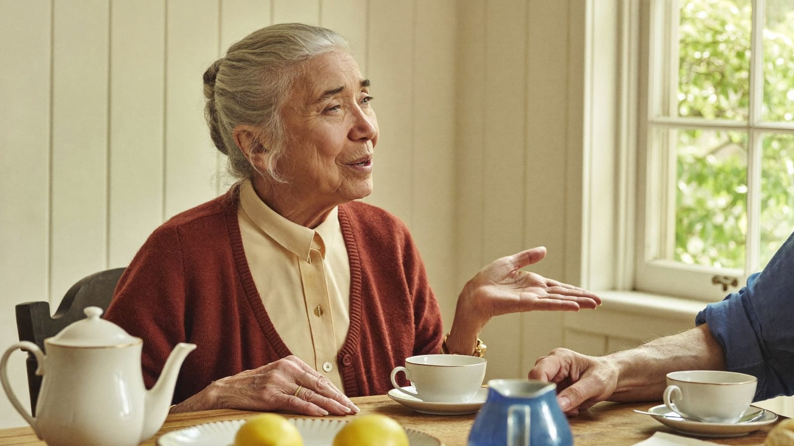 An older woman at her kitchen table in morning light, caught mid-sentence with one hand lifted, talking to someone out of frame.