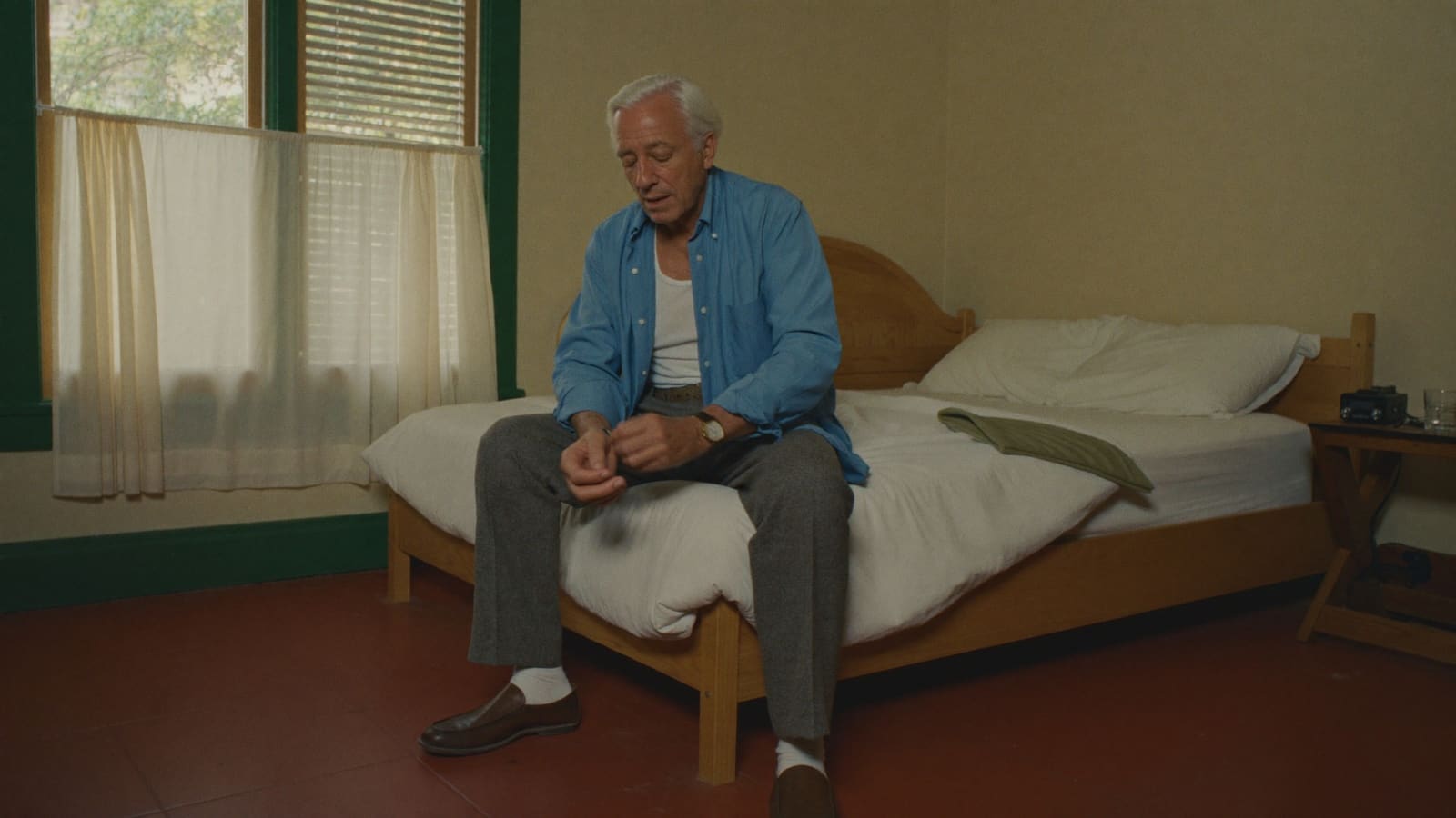 An older man sitting on the edge of his own bed, buttoning his shirt cuff in the morning light.