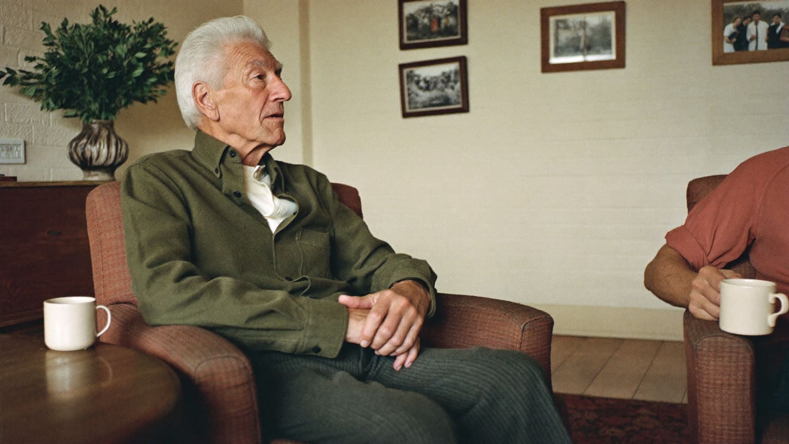 An older man in an armchair talking with a visitor seated opposite him in a suburban lounge room.