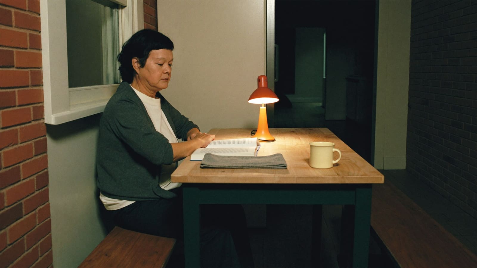 A support worker sitting alone at a kitchen table late at night, reading by the light of a single lamp.