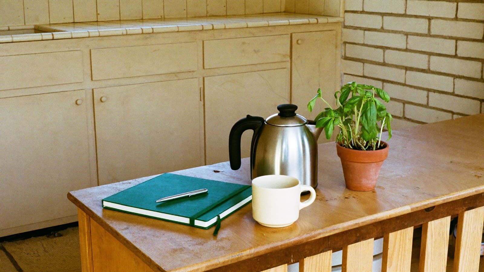 A closed green hardcover notebook with a slim silver pen resting on top, on a honey-toned timber kitchen bench beside a plain cream mug, a stainless steel kettle and a small terracotta pot of basil.