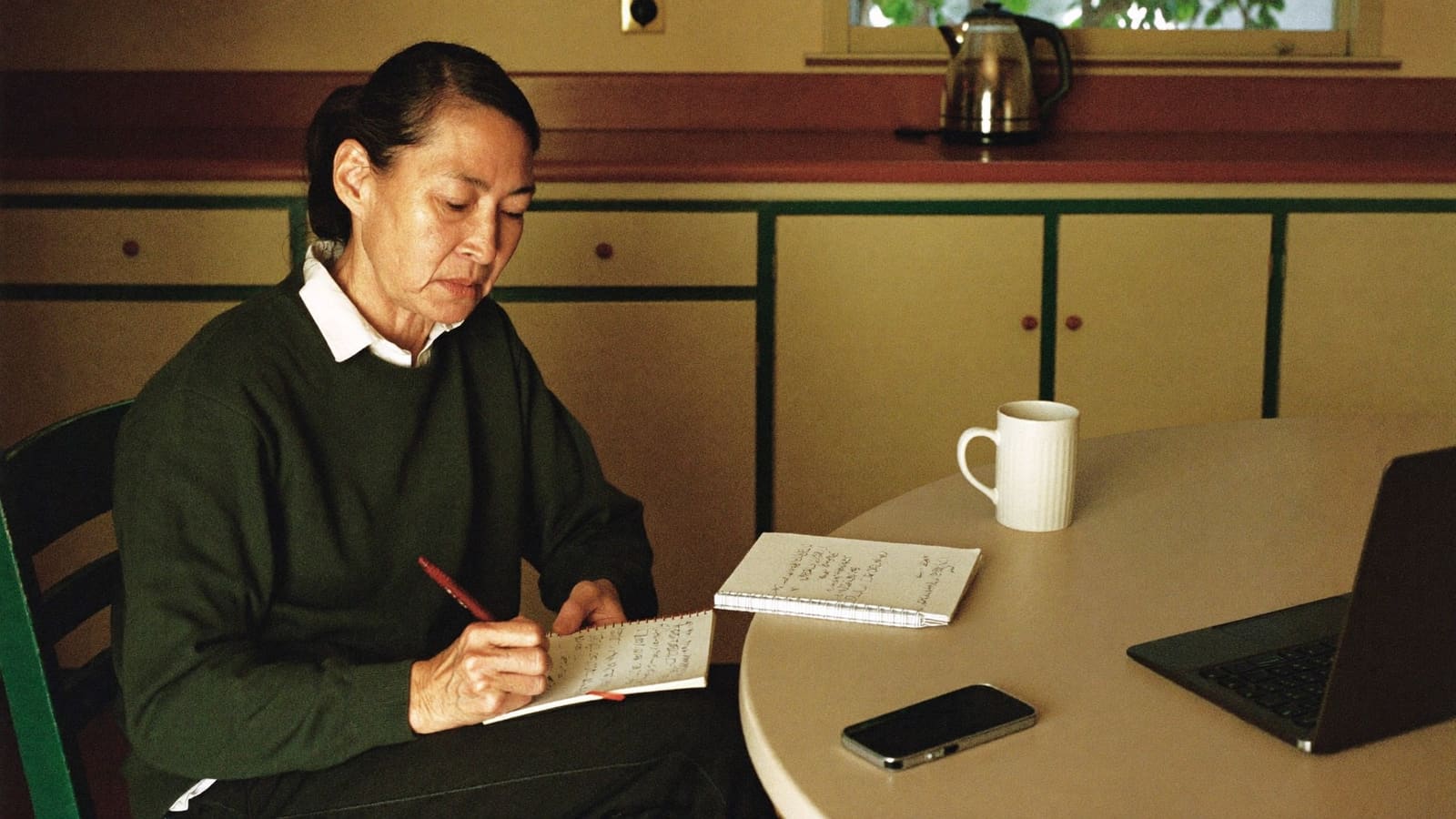A woman writing a short list in a notebook at her parent's kitchen table in the late afternoon.