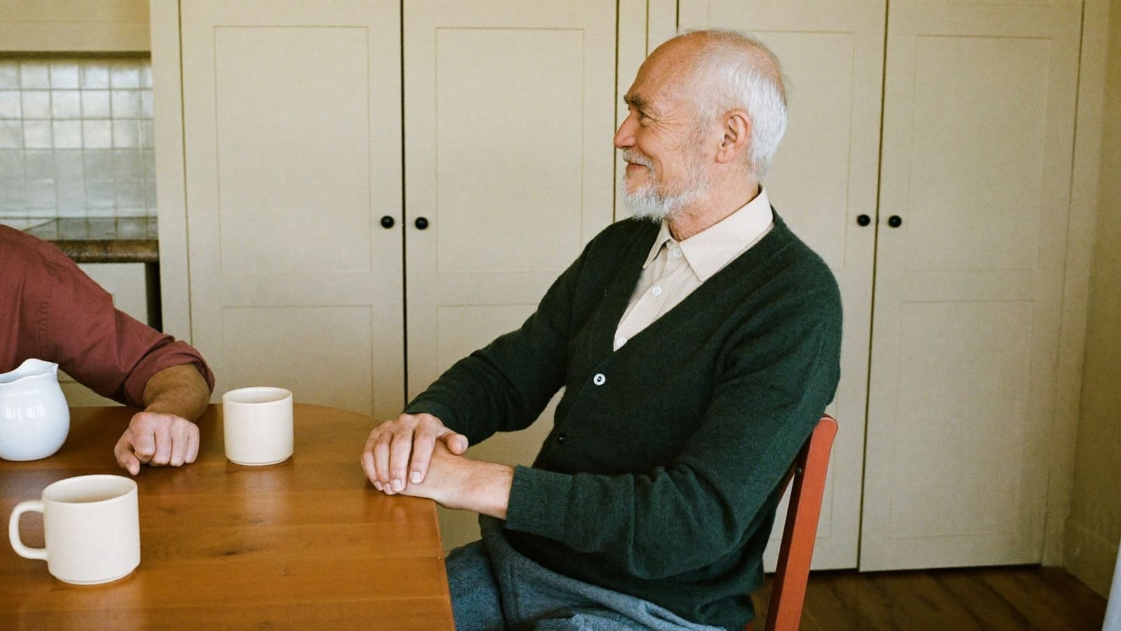 An older man with white hair and a white beard in a dark green cardigan, sitting in side profile at a timber kitchen table with his hands clasped, smiling at someone whose forearm in a deep red rolled sleeve rests on the table at the edge of the frame.