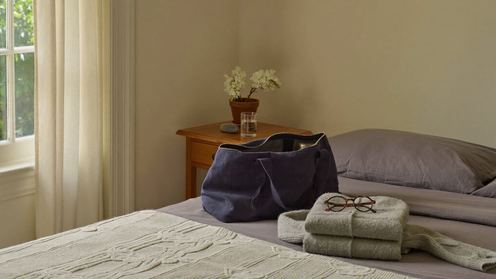 A small navy overnight bag open on a neatly made bed beside a folded grey cardigan and a pair of reading glasses.