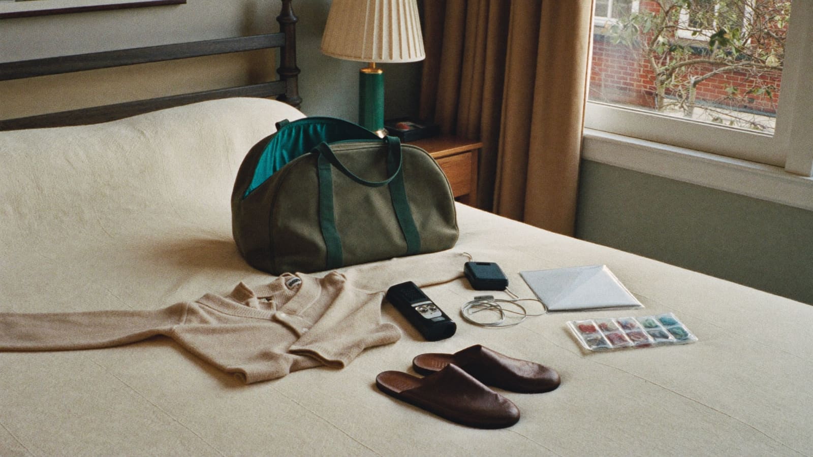An olive canvas bag open on a cream bedspread, with a cardigan, torch, small radio, pill organiser, document sleeve and slippers laid out beside it.